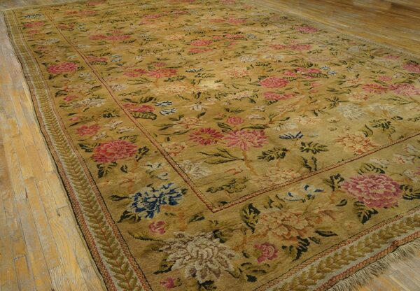 Large gold floral rug with pink and blue motifs features a fringed edge on light wood planks.