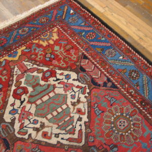 Corner of a low-pile red rug with blue borders and complex traditional patterns on light wood flooring.