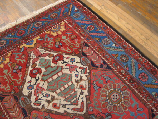 Corner of a low-pile red rug with blue borders and complex traditional patterns on light wood flooring.