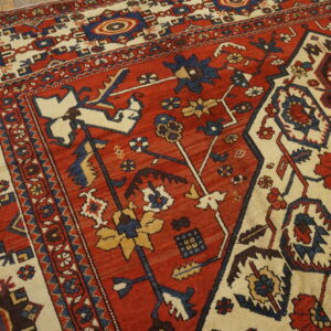 Traditional patterned rug with a red field, cream border, and stylized motifs resting on wooden flooring.
