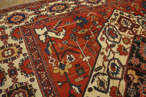 Traditional patterned rug with a red field, cream border, and stylized motifs resting on wooden flooring.