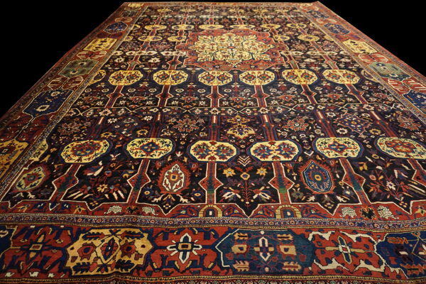 Dark area rug featuring complex traditional patterns in navy, red, and gold, angled on a black background.