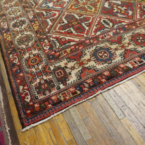 Low-pile red, cream, and blue geometric rug corner on rustic hardwood floors with rolled rugs nearby.