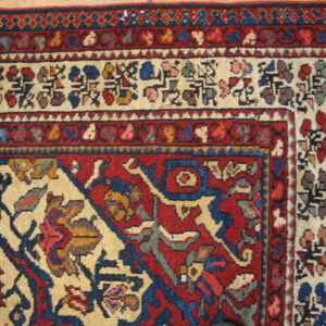 A low-pile rug corner featuring complex red, cream, and blue traditional patterns, resting on light wood flooring.