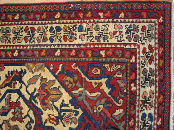 A low-pile rug corner featuring complex red, cream, and blue traditional patterns, resting on light wood flooring.