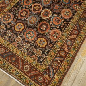 A detailed view of a dark floral patterned rug with rust and blue motifs placed on light wooden floors.