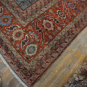 Red and blue patterned low-pile rug on distressed wood floor next to a rolled rug and a gray shoe.