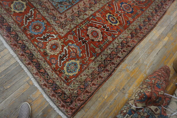 Red and blue patterned low-pile rug on distressed wood floor next to a rolled rug and a gray shoe.