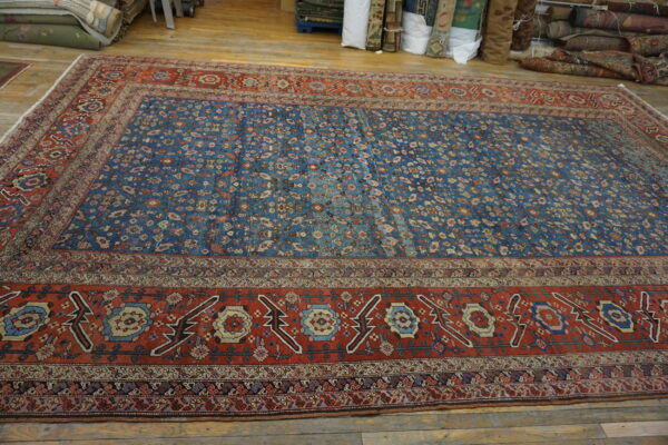 A large rectangular rug with a low blue field and complex red-orange traditional borders on wood planks.