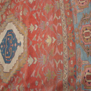 Muted red rug close-up with geometric blue and tan stylized patterns surrounding a large cream stepped design.