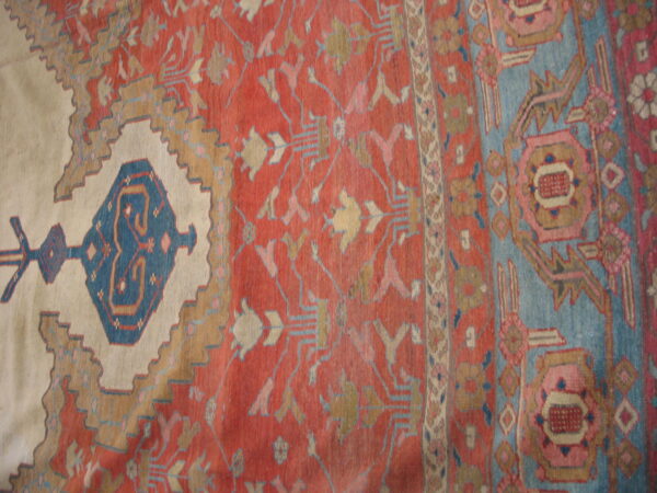 Muted red rug close-up with geometric blue and tan stylized patterns surrounding a large cream stepped design.