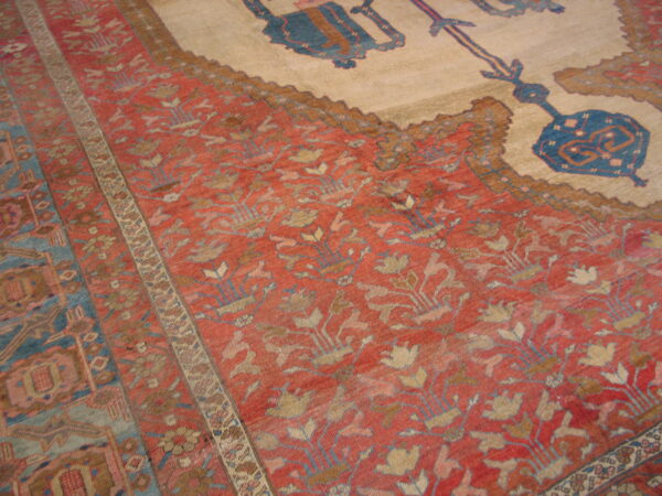 Close view of a rug's red floral field, blue patterned borders, and a large cream center with blue motifs.
