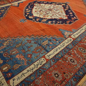 A reddish-orange rug with large blue panels, ornate patterns, and borders containing script on wood floors.