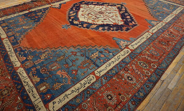 A reddish-orange rug with large blue panels, ornate patterns, and borders containing script on wood floors.