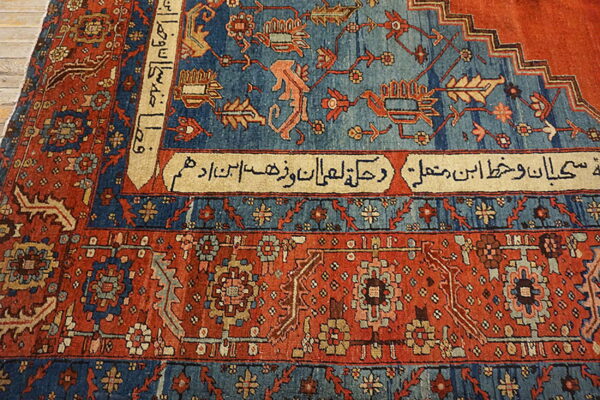 Red and blue patterned rug showing script cartouches and animal motifs rests on light wood plank flooring.