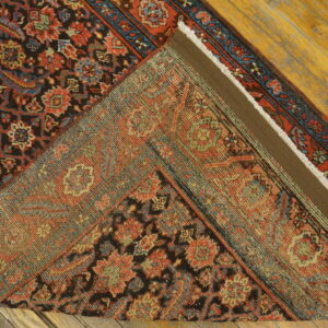 Close-up of a low-pile red, black, and blue patterned rug folded back on distressed wood flooring.