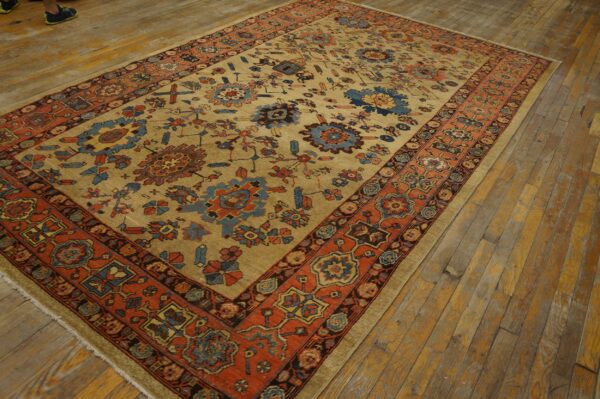 Tan rug featuring rust orange, blue, and gold floral patterns and an ornate border on distressed wood flooring.