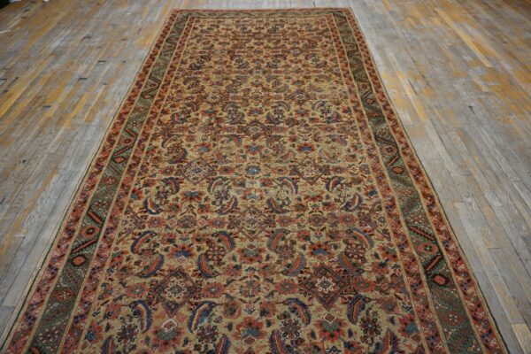 A long runner rug featuring a dense tan, red, and blue traditional pattern on worn wood flooring.