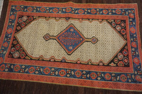 A textured rug with a central blue diamond medallion on a beige field and geometric red-orange borders.