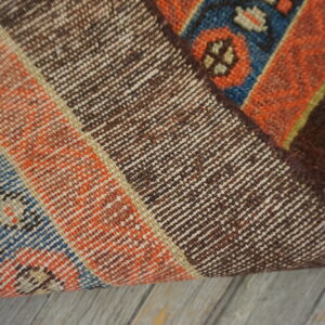 Rug corner folded back exposing the tight brown weave backing next to the low-pile orange and blue pattern.