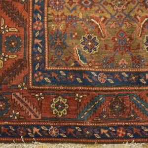 Red, navy, and olive traditional rug corner with fringe displayed on light wood floorboards.