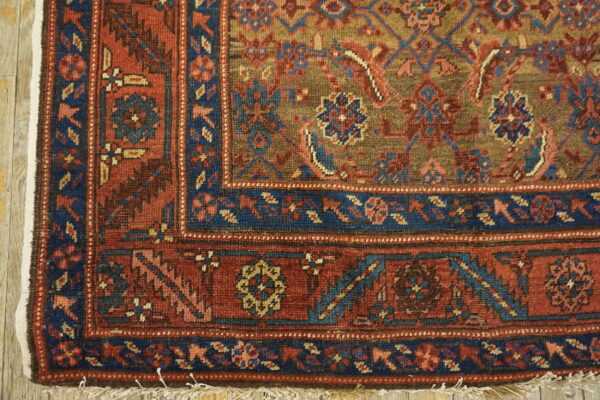 Red, navy, and olive traditional rug corner with fringe displayed on light wood floorboards.