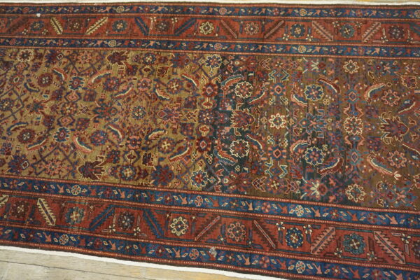 Traditional low-pile rug with geometric and floral patterns in red, khaki, and blue, visible on wood planks.