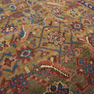 Textured rug close-up showing traditional motifs detailed in blue, rose, and maroon on an olive field.