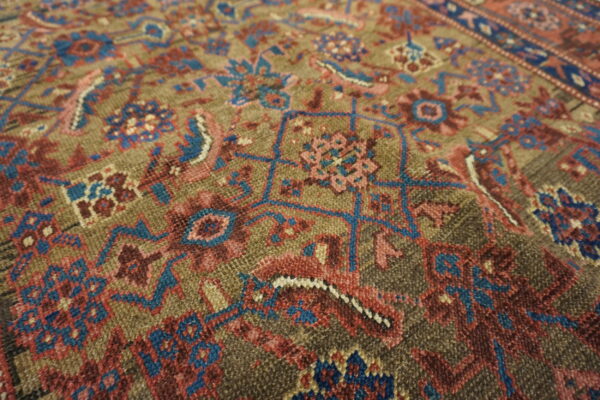 Textured rug close-up showing traditional motifs detailed in blue, rose, and maroon on an olive field.