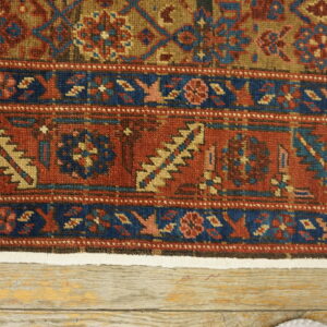 Detailed low-pile rug border in rust, navy blue, and tan lying on light distressed wood floorboards.