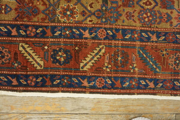 Detailed low-pile rug border in rust, navy blue, and tan lying on light distressed wood floorboards.