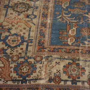 Distressed traditional rug corner showing blue, beige, and orange motifs next to yellow wood floor.