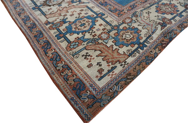 Corner detail of a traditional patterned rug in beige, blue, and rust on a white studio background.