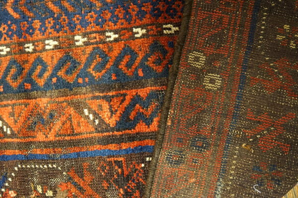 Closeup of a folded rug displays orange and blue pile patterns contrasting with the dark brown reverse side.
