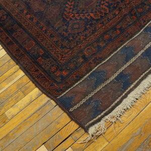 Muted blue and red traditional rug corner with decorative fringe rests on aged yellow wooden floorboards.