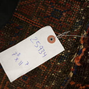 A white retail tag obscures part of a dark, low-pile rug featuring red, blue, and cream geometric designs.