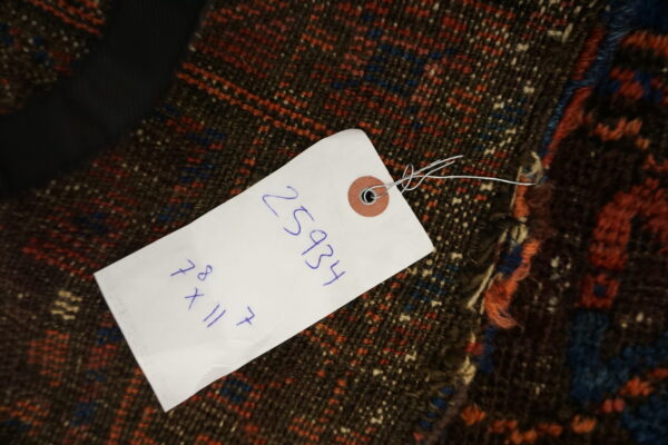 A white retail tag obscures part of a dark, low-pile rug featuring red, blue, and cream geometric designs.