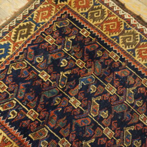 Geometric tribal rug in deep navy, red, gold, and brown colors rests on weathered light wood plank flooring.