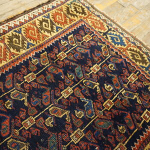 Low pile rug corner with navy, red, and gold geometric patterns rests on light wood plank flooring.