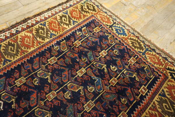 Low pile rug corner with navy, red, and gold geometric patterns rests on light wood plank flooring.