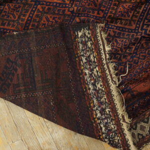 Dark red and navy geometric rug folded on light wood planks, revealing the flat woven backing and fringe.