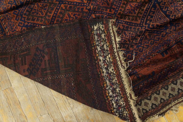 Dark red and navy geometric rug folded on light wood planks, revealing the flat woven backing and fringe.
