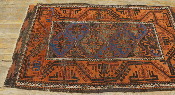 Worn, low-pile rug featuring bold rust-orange and dark blue geometric patterns on light wood.