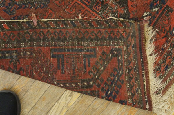Red and dark patterned rug folded over light wood flooring, revealing tight weaving and fringe.