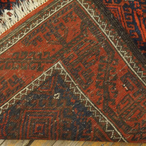 Close-up of a folded red and navy geometric rug with fringe, lying on wooden floorboards.