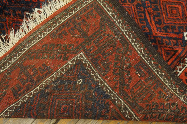 Close-up of a folded red and navy geometric rug with fringe, lying on wooden floorboards.