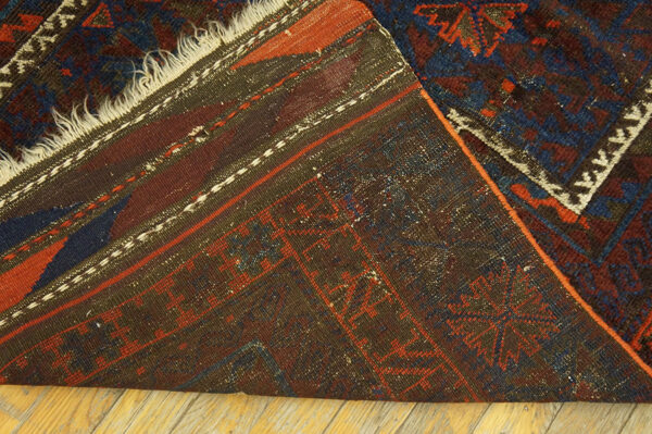 A dark red and navy patterned rug is folded on a wood floor, exposing the woven backing.