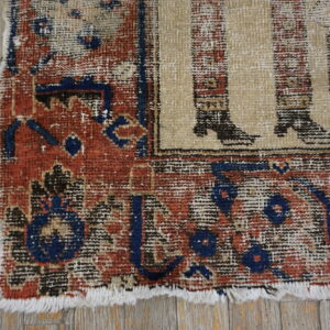 Worn corner of a rug showing red and blue borders, a beige figurative pattern, and fringe on wood planks.