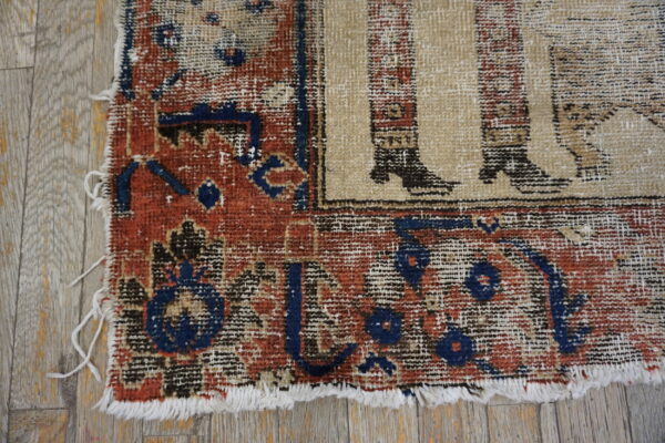 Worn corner of a rug showing red and blue borders, a beige figurative pattern, and fringe on wood planks.