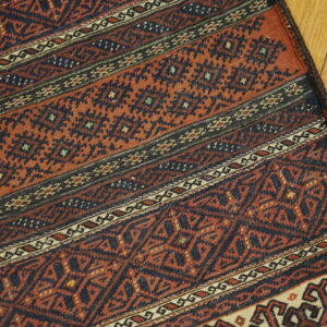 Terracotta and navy rug with repeating geometric banded patterns rests on light hardwood flooring.